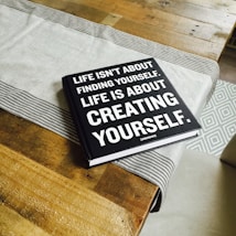 A black hardcover book with white text on the cover is placed on a wooden table. A light gray table runner with striped edges lies underneath the book. The book's cover reads, 'Life isn't about finding yourself. Life is about creating yourself.' The image also features the corner of a patterned rug in the background.