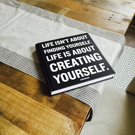 A black hardcover book with white text on the cover is placed on a wooden table. A light gray table runner with striped edges lies underneath the book. The book's cover reads, 'Life isn't about finding yourself. Life is about creating yourself.' The image also features the corner of a patterned rug in the background.