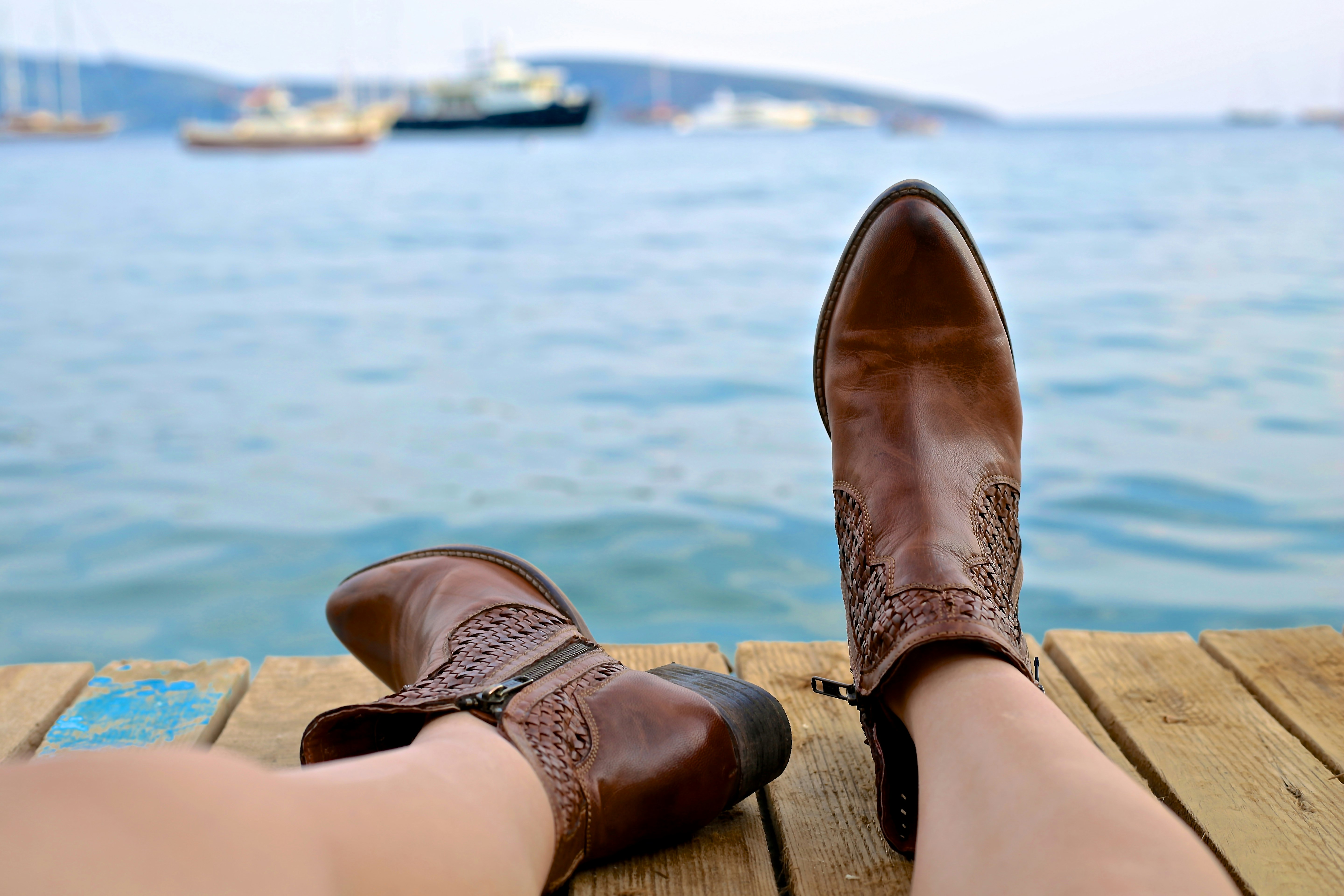 person wearing brown leather side-zip boots sits on brown wooden pier near body of water during daytime