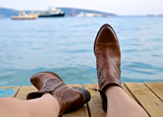 person wearing brown leather side-zip boots sits on brown wooden pier near body of water during daytime