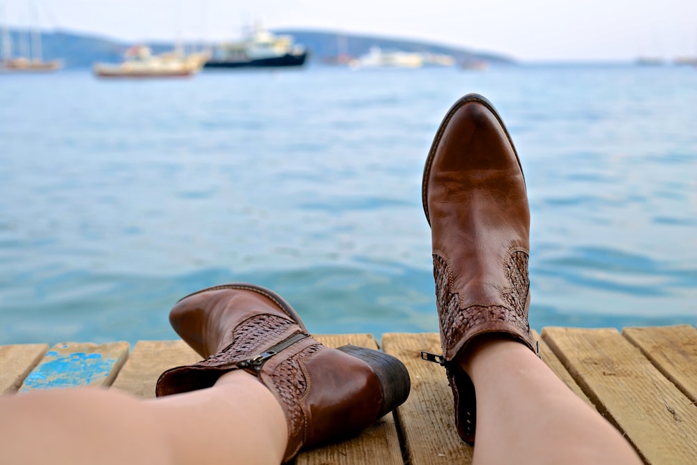 person wearing brown leather side-zip boots sits on brown wooden pier near body of water during daytime