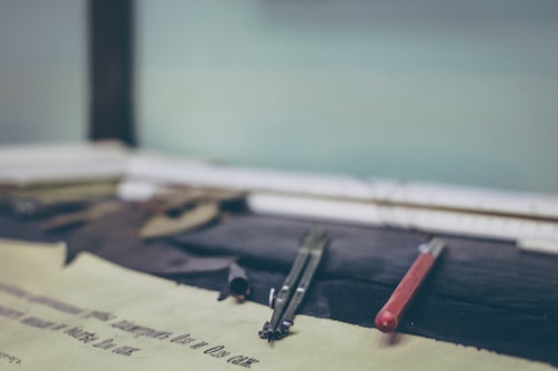 A close-up of drafting tools including a compass and a red mechanical pencil on a desk with blurred papers in the background. The focus is on the tools, suggesting a scene of technical drawing or drafting.