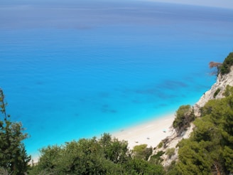 A serene coastal landscape featuring a vibrant turquoise sea meeting a sandy beach. The coastline is lined with rugged cliffs and lush greenery. Sparse beach umbrellas are visible on the sand, and the calm water stretches out to the horizon.