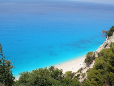 A serene coastal landscape featuring a vibrant turquoise sea meeting a sandy beach. The coastline is lined with rugged cliffs and lush greenery. Sparse beach umbrellas are visible on the sand, and the calm water stretches out to the horizon.