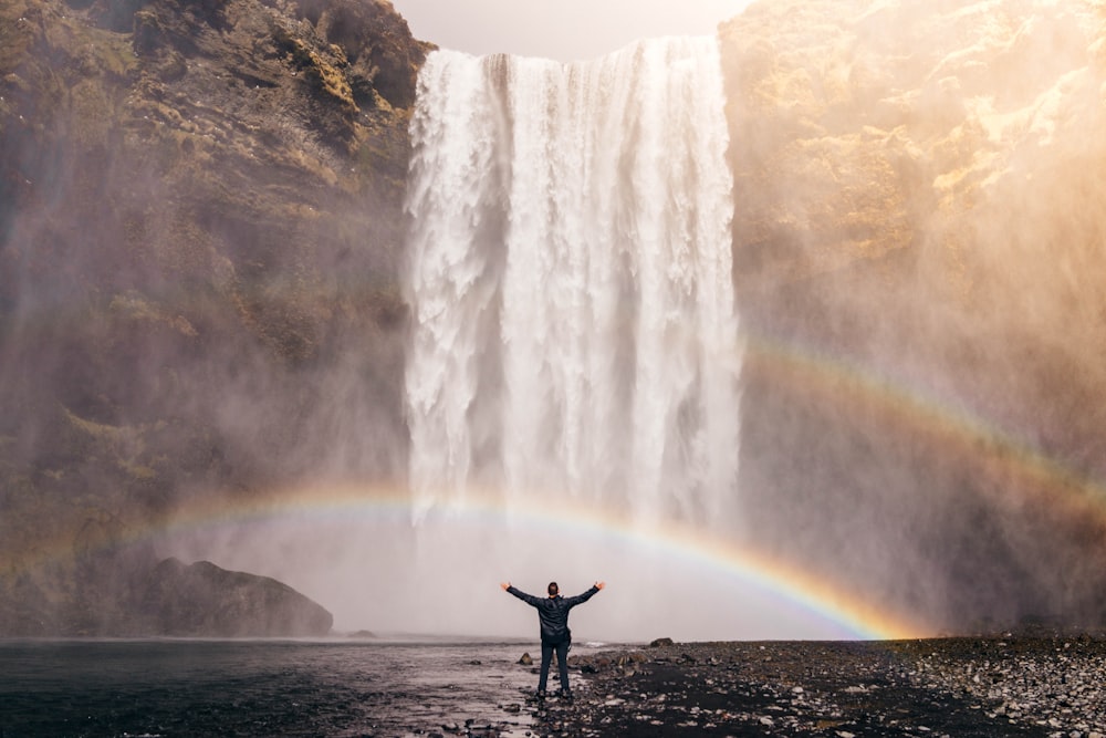 person in front of waterfalls with double rainbow during daytime