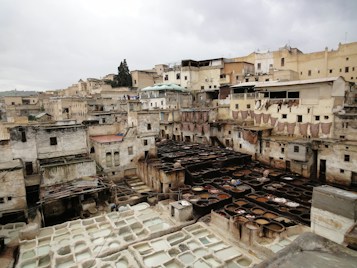 An array of circular, stone-lined pits are filled with various liquids in a traditional leather tannery. These tanneries are surrounded by old, weathered buildings made of stone and bricks, with some rooftops visible. Pieces of animal hides are draped on the walls and rooftops to dry, and the scene looks busy and industrial.