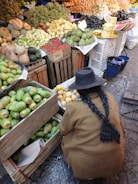 A market scene with a wide variety of fruits, including mangoes, pineapples, and grapes neatly arranged in wooden crates. A person with long braided hair and wearing a brown coat is crouched down, possibly selecting or arranging fruit.