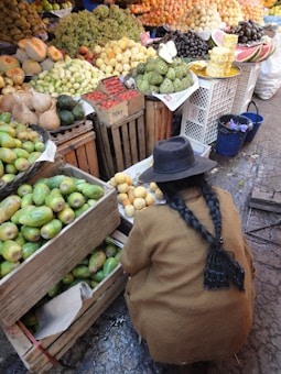 A market scene with a wide variety of fruits, including mangoes, pineapples, and grapes neatly arranged in wooden crates. A person with long braided hair and wearing a brown coat is crouched down, possibly selecting or arranging fruit.