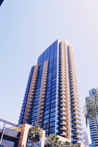 A modern high-rise building with numerous balconies, characterized by a blue-tinted glass facade and a sleek architectural design. Palm trees are visible at the base of the building in an urban setting, under a clear blue sky.