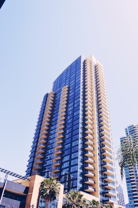 A modern high-rise building with numerous balconies, characterized by a blue-tinted glass facade and a sleek architectural design. Palm trees are visible at the base of the building in an urban setting, under a clear blue sky.