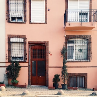 A peach-colored building facade featuring three windows with brown shutters and metal bars. A central ornate wooden door is surrounded by brick accents. Potted evergreen plants are placed on either side of the door, adding a touch of greenery to the paved sidewalk.