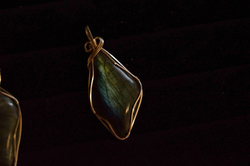A pendant featuring a polished stone with a glossy surface, encased in intricately wrapped metal wire. The stone exhibits hues of blue and green with subtle iridescence. The wire wrapping includes a loop at the top for attachment.