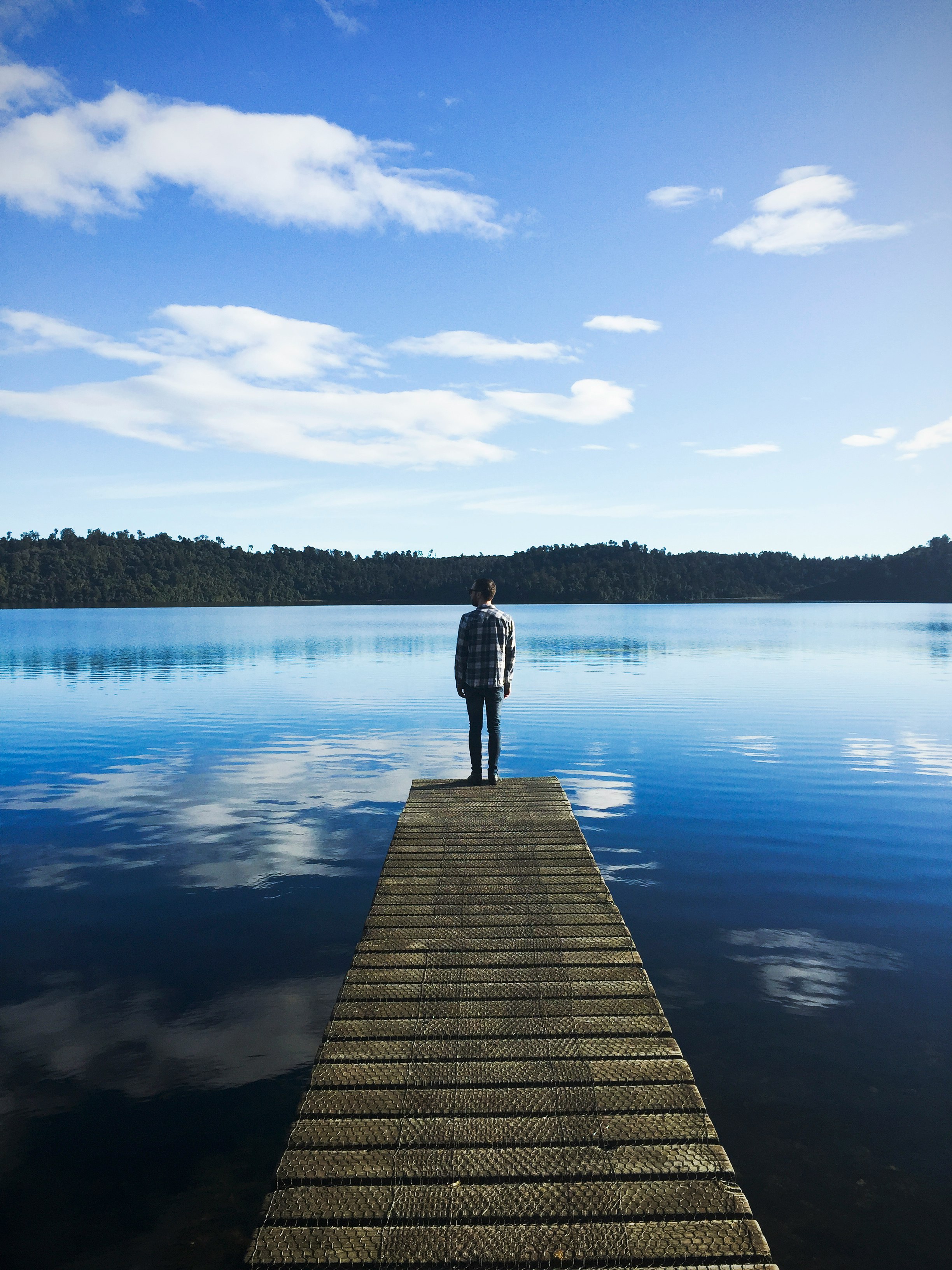 man standing on lake dock watching water under blue sky during daytime