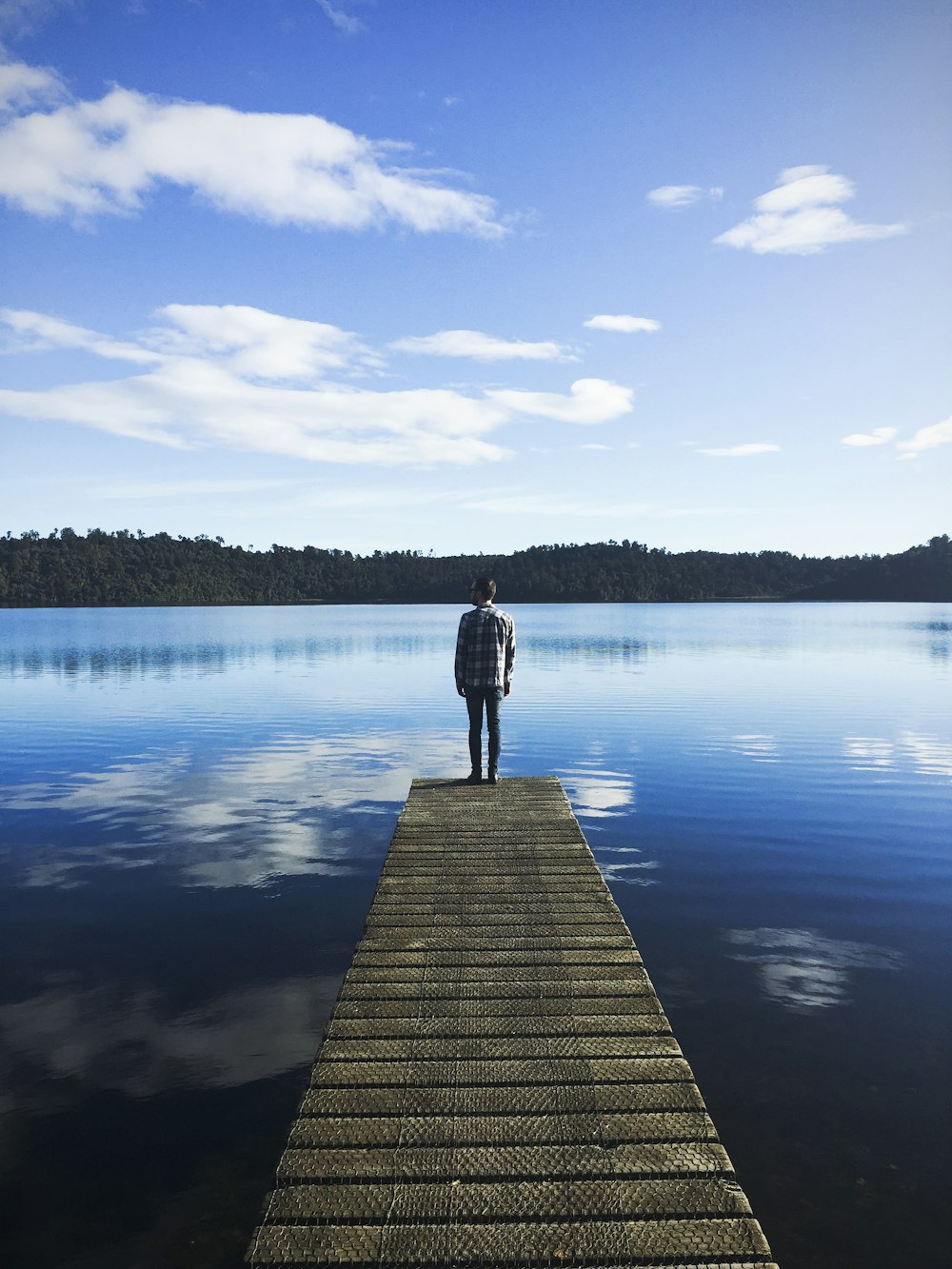 uomo in piedi sul molo del lago che guarda l'acqua sotto il cielo blu durante il giorno