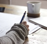 person writing on brown wooden table near white ceramic mug