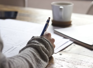 person writing on brown wooden table near white ceramic mug