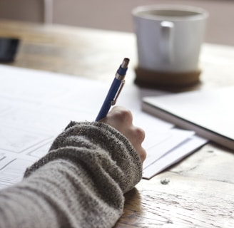 person writing on brown wooden table near white ceramic mug