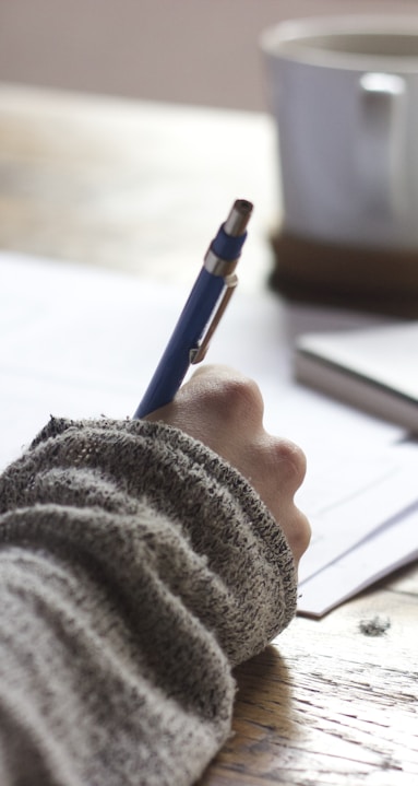 person writing on brown wooden table near white ceramic mug