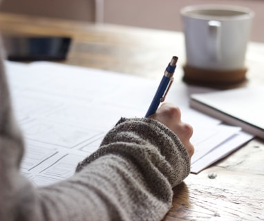 person writing on brown wooden table near white ceramic mug