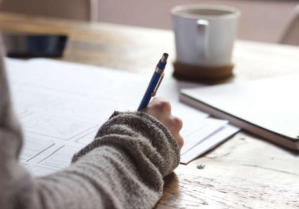 person writing on brown wooden table near white ceramic mug