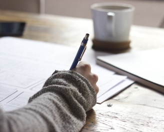 person writing on brown wooden table near white ceramic mug