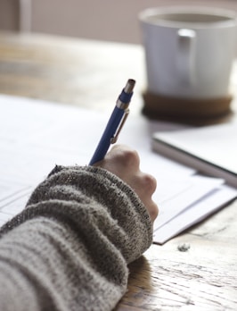 person writing on brown wooden table near white ceramic mug