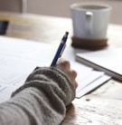 person writing on brown wooden table near white ceramic mug