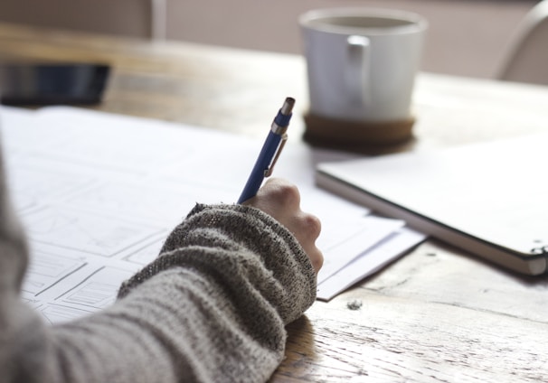 person writing on brown wooden table near white ceramic mug