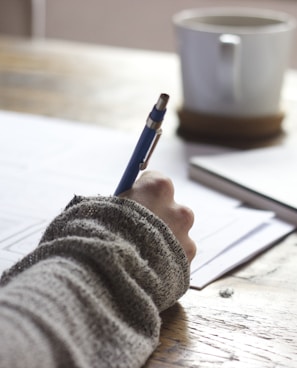 person writing on brown wooden table near white ceramic mug