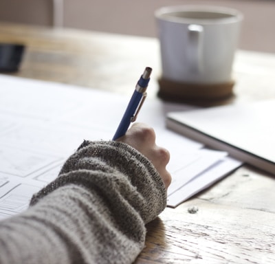 person writing on brown wooden table near white ceramic mug