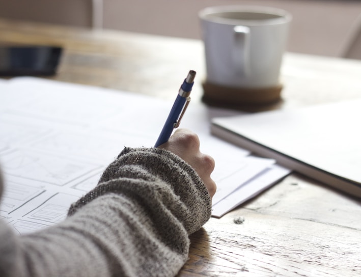 person writing on brown wooden table near white ceramic mug