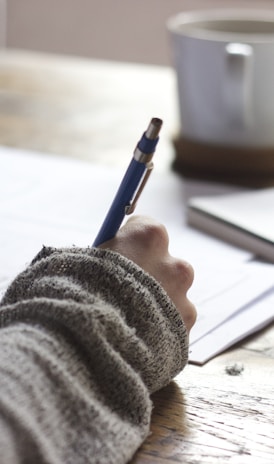 person writing on brown wooden table near white ceramic mug