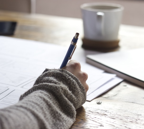 person writing on brown wooden table near white ceramic mug