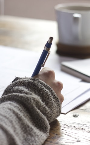 person writing on brown wooden table near white ceramic mug