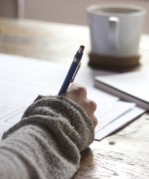 person writing on brown wooden table near white ceramic mug