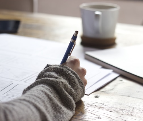 person writing on brown wooden table near white ceramic mug