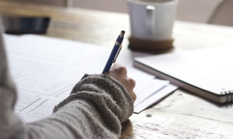 person writing on brown wooden table near white ceramic mug