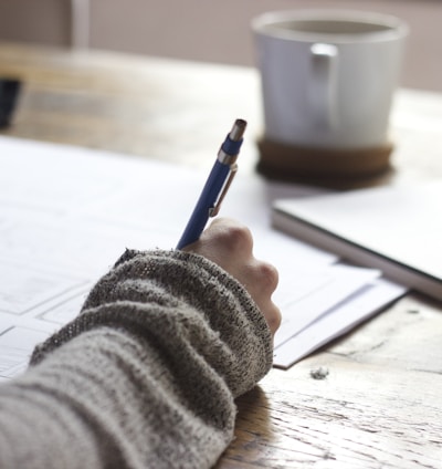 person writing on brown wooden table near white ceramic mug