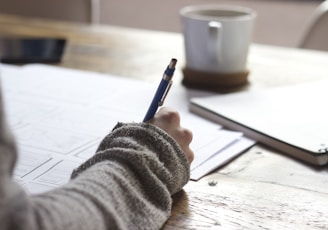 person writing on brown wooden table near white ceramic mug