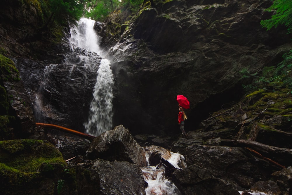 person standing on rock formation under red umbrella beside waterfalls