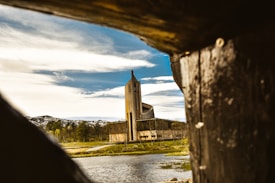 A modern, uniquely designed church is seen through the opening of a rustic wooden structure. A serene landscape surrounds the building with a body of water in the foreground and hills with sparse trees in the background under a partly cloudy sky.
