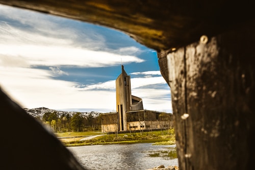 A modern, uniquely designed church is seen through the opening of a rustic wooden structure. A serene landscape surrounds the building with a body of water in the foreground and hills with sparse trees in the background under a partly cloudy sky.