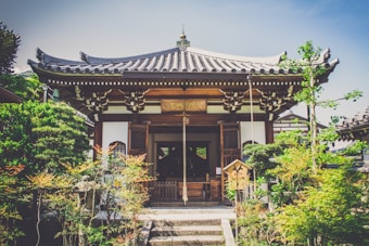 A traditional Japanese wooden building with a curved tile roof surrounded by lush green plants and small trees. The structure is ornately decorated with woodwork and features a sign with Japanese characters above the entrance.