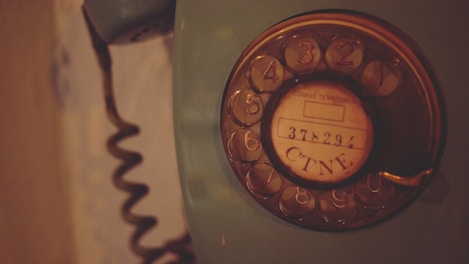 A vintage rotary dial telephone with a coiled cord is seen. The dial has numbers from 1 to 0 and a central circle with the text '378291 CTNE'. The phone is an old-fashioned device with a dark, muted color palette.
