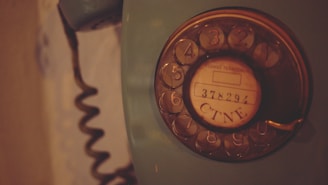 A vintage rotary dial telephone with a coiled cord is seen. The dial has numbers from 1 to 0 and a central circle with the text '378291 CTNE'. The phone is an old-fashioned device with a dark, muted color palette.