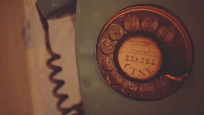 A vintage rotary dial telephone with a coiled cord is seen. The dial has numbers from 1 to 0 and a central circle with the text '378291 CTNE'. The phone is an old-fashioned device with a dark, muted color palette.