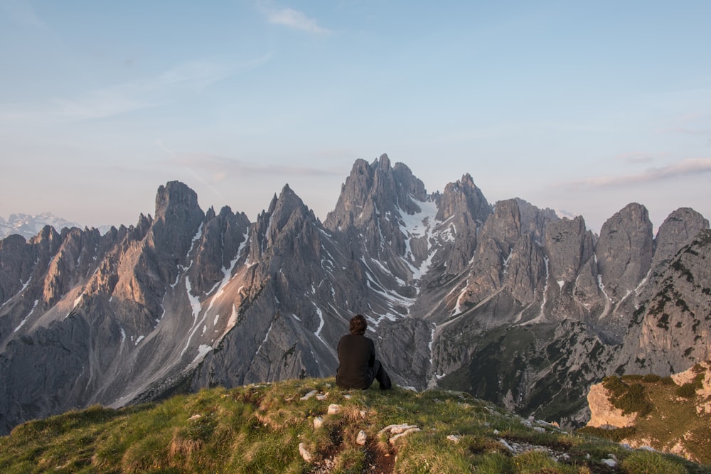 man sitting on cliff facing gray mountain