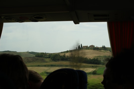 The photograph captures a scenic countryside landscape viewed from inside a vehicle. Rolling hills with neatly cultivated fields stretch into the distance, adorned with scattered trees and a cluster of buildings on a hilltop. Red curtains frame the window, and the silhouette of passengers is visible, creating a peaceful and contemplative mood.