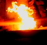 Bright orange flames with intense heat illuminate a dark setting. The fire appears to be emerging from a ground surface, creating a stark contrast with the surrounding shadows.