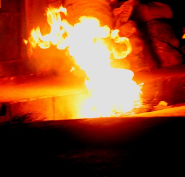 Bright orange flames with intense heat illuminate a dark setting. The fire appears to be emerging from a ground surface, creating a stark contrast with the surrounding shadows.